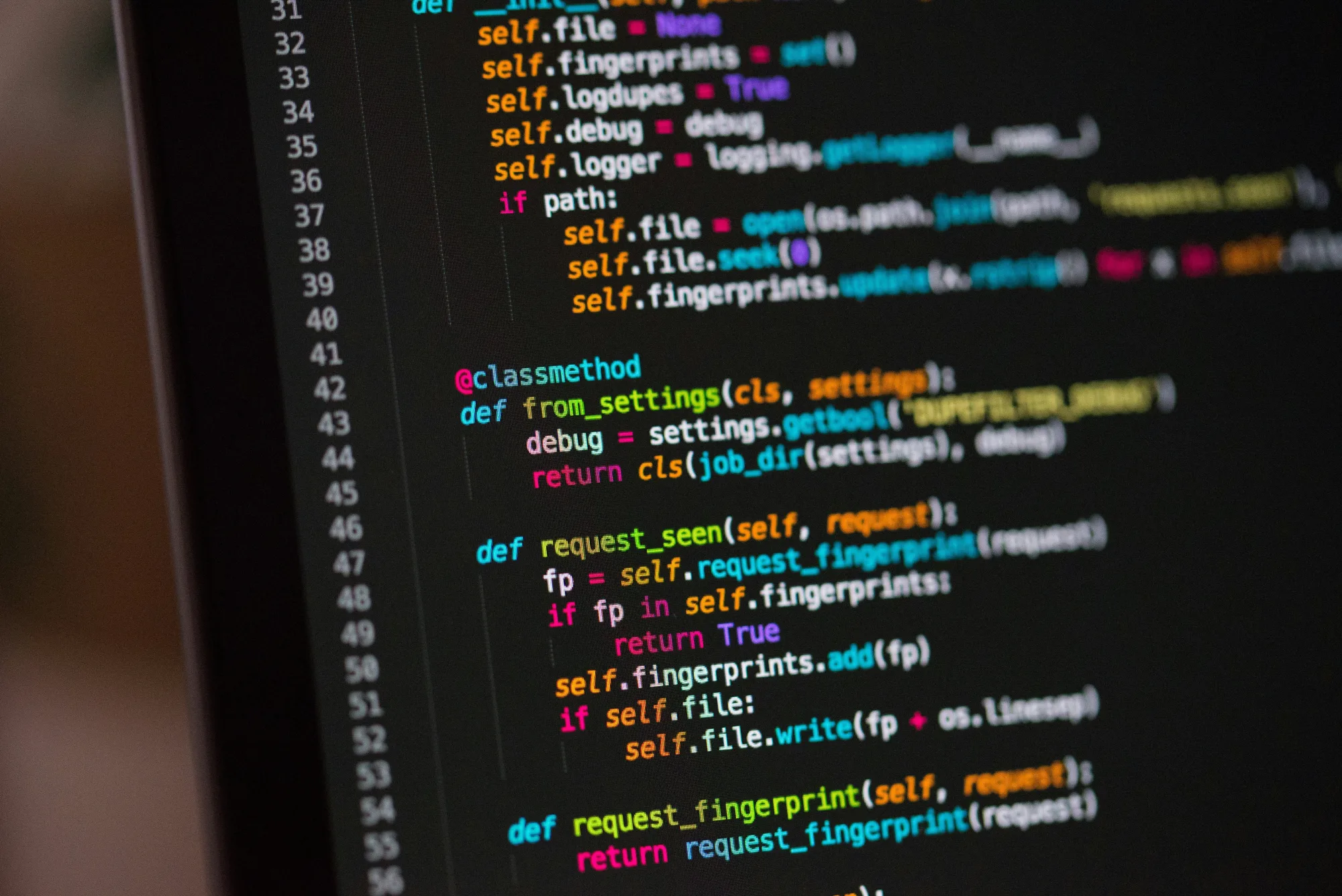A close-up photo of a monitor showing Python code in a dark editor theme, lines 31 to 56, sharp on the left and blurring toward the right.